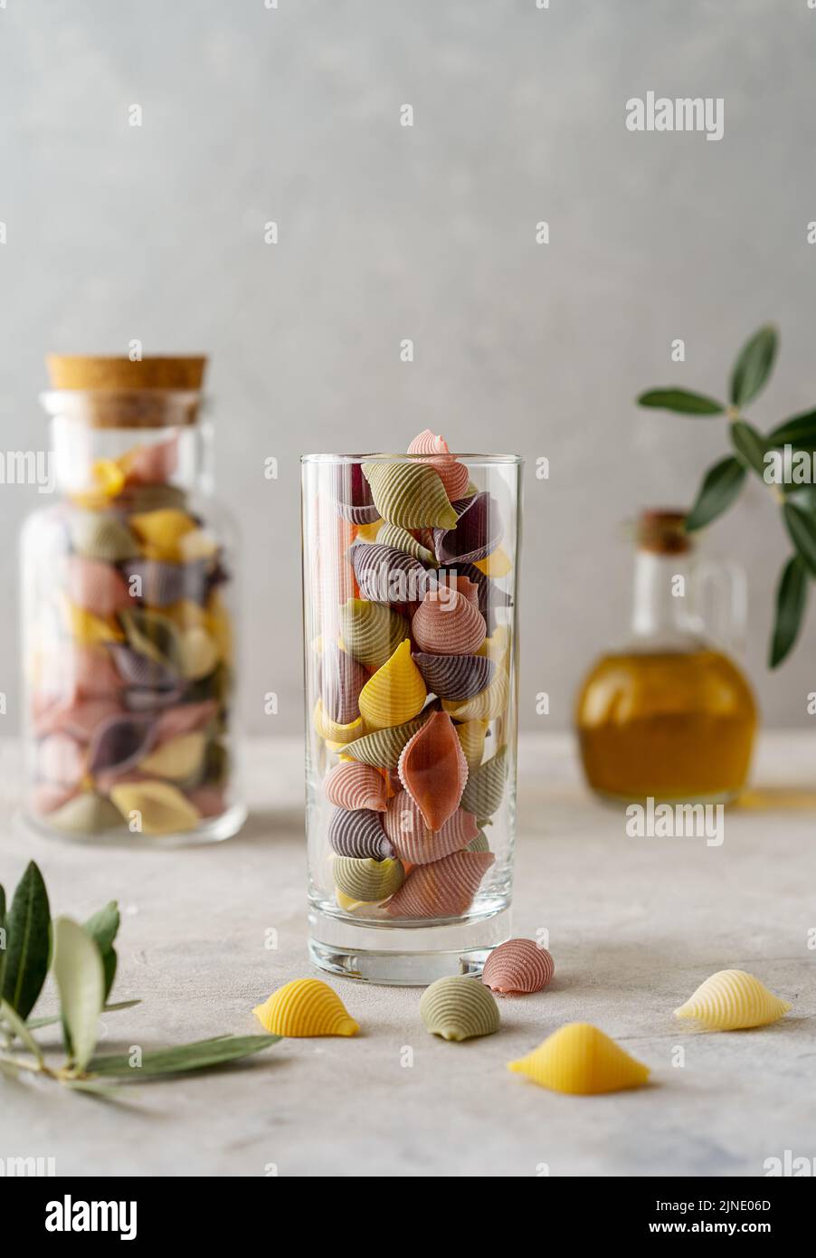 Mehrfarbige italienische Pasta Conchiglie oder Muscheln in Glas und Flasche, Olivenöl, Olivenzweig. Konkreter Hintergrund Stockfoto