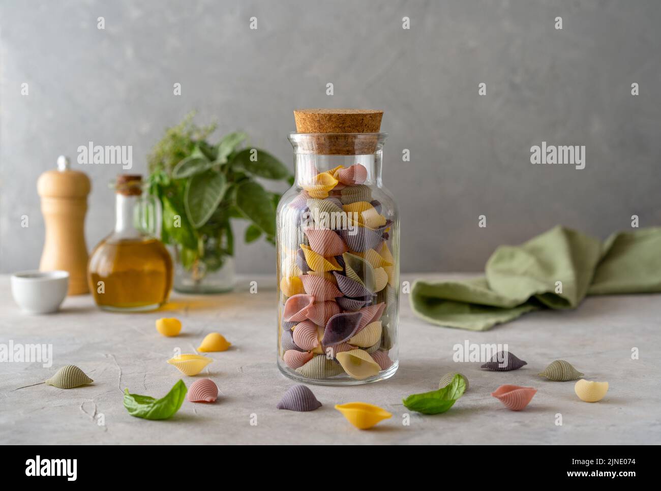 Mehrfarbige italienische Pasta Conchiglie oder Muscheln in Glasflasche, Olivenöl, Olivenzweig. Konkreter Hintergrund Stockfoto