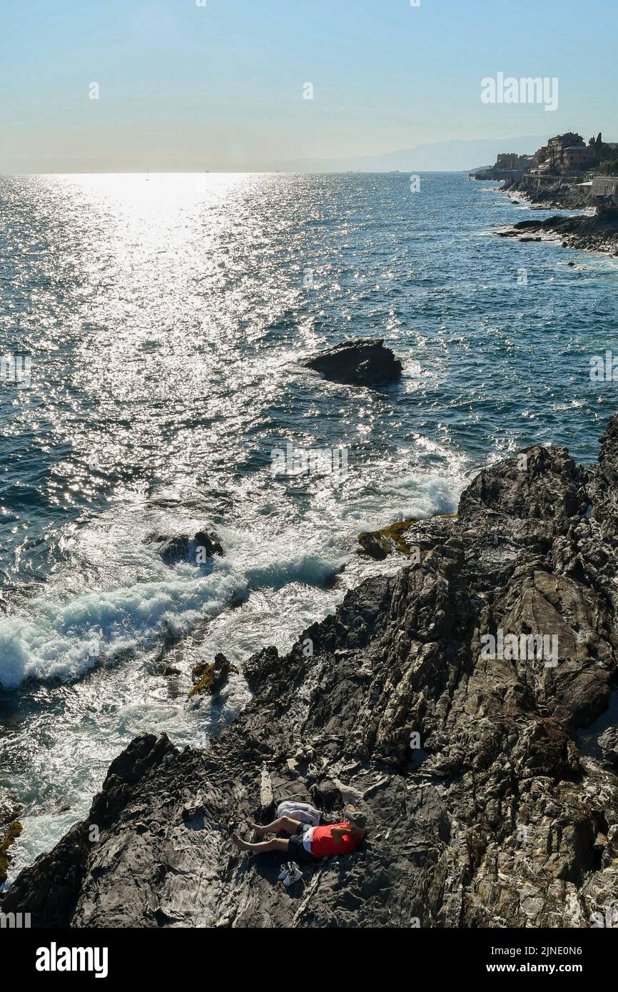 Blick auf den Golf des Paradieses mit einem Mann, der auf den Felsen am Meer liegt, und dem Fischerdorf im Hintergrund, Nervi, Genua, Ligurien, Italien Stockfoto