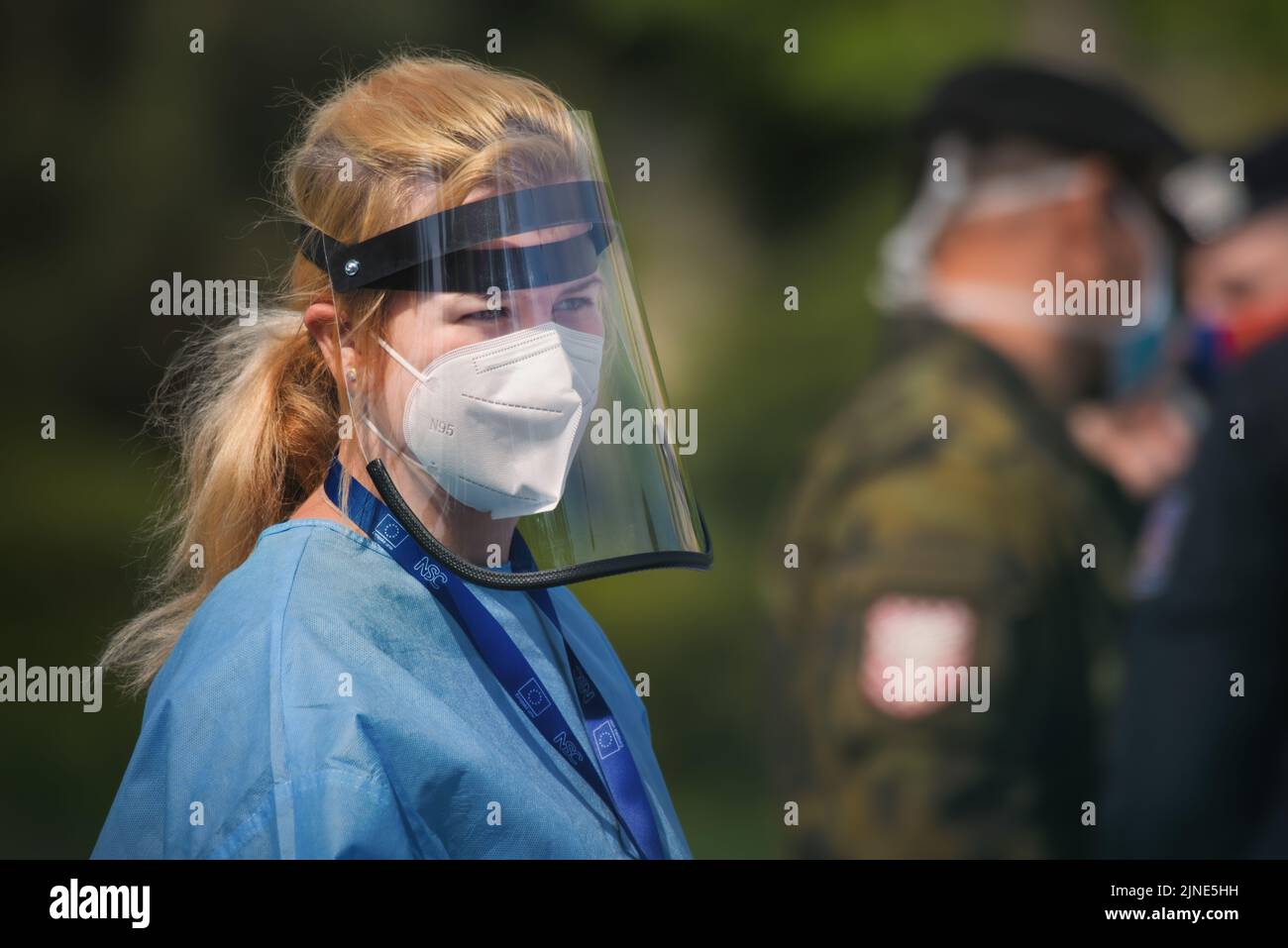 Medic trägt ein Atemschutzgerät und einen Gesichtsschutz aus Kunststoff. Hinter ihr sind Mitglieder der Militärpolizei, die Leute überprüfen, die auf einen Corona-Virus-Test warten. Stockfoto
