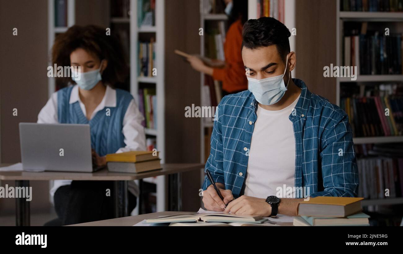 Studenten studieren in der Bibliothek konzentriert intelligente junge indische Kerl in Schutzmaske sitzt am Schreibtisch Hausaufgaben Vorbereitung für die Prüfung schreibt Notizen in Stockfoto