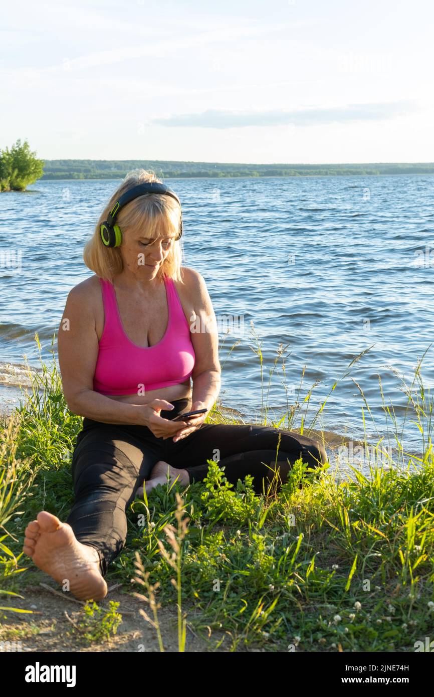 Eine ältere Frau in Sportbekleidung und Kopfhörern wählt Musik für den Sport, sieht Online-Übungen für das Coaching in einem Park am Ufer eines Sees, die se Stockfoto