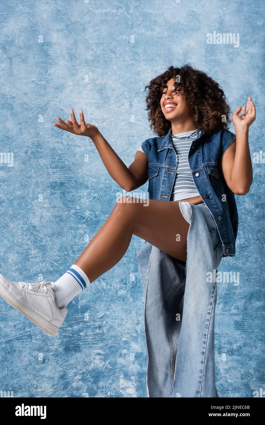 Glückliche afroamerikanische Frau in zerrissenen Jeans, die auf einem Bein steht und mit der Hand auf blauen Hintergrund zeigt Stockfoto