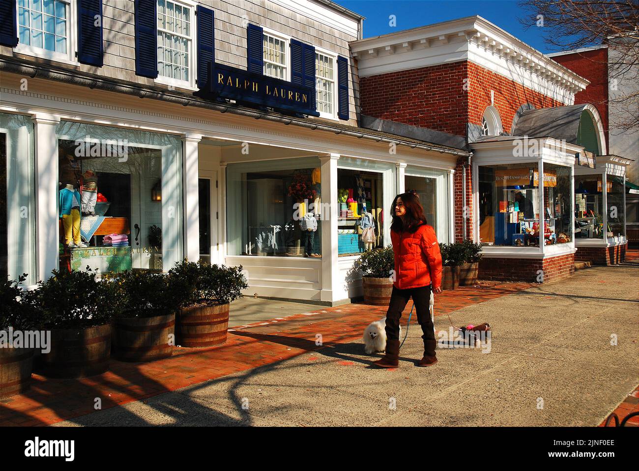 Eine junge Frau geht mit ihren Hunden an den luxuriösen Designerläden in East Hampton, Long Island, New York, einer der wohlhabenden Städte der Hamptons, vorbei Stockfoto