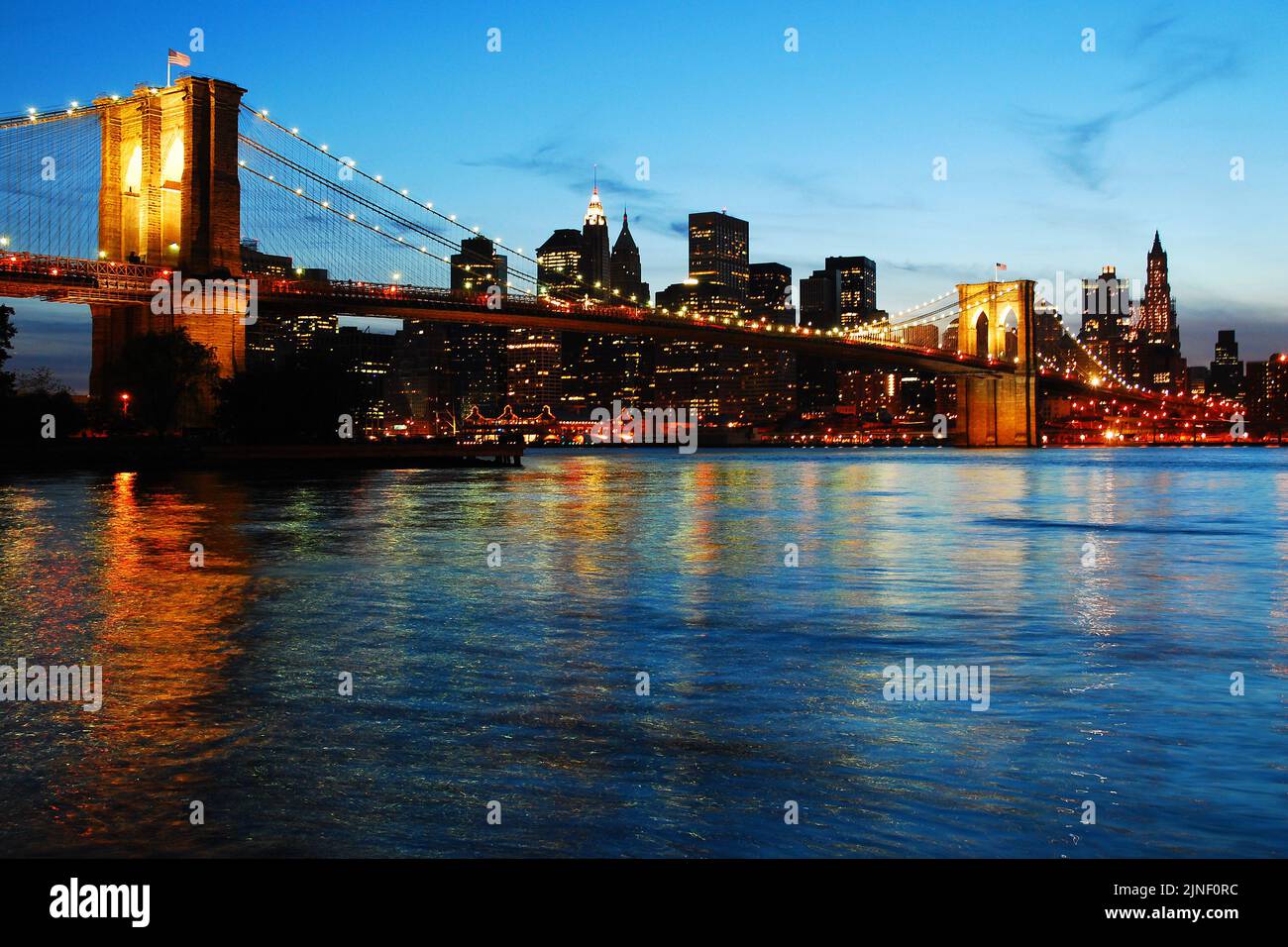 Die Beleuchtung der Brooklyn Bridge spiegelt sich im Wasser des East River wider, wenn sich die Dämmerung auf der Skyline von Manhattan nähert Stockfoto