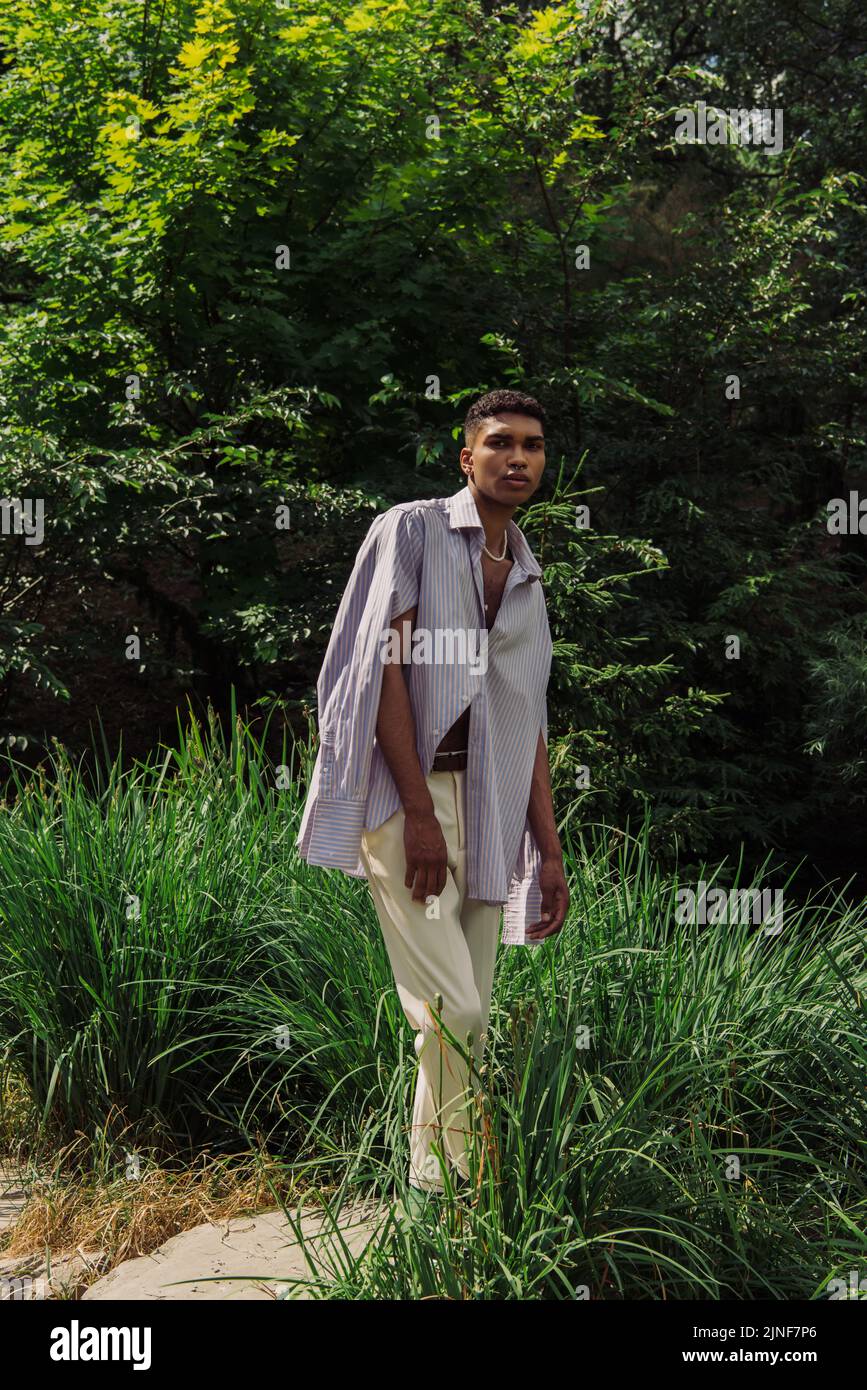 afroamerikanischer Mann in trendigem Hemd und weißer Hose beim Blick auf die Kamera im grünen Park, Stockbild Stockfoto