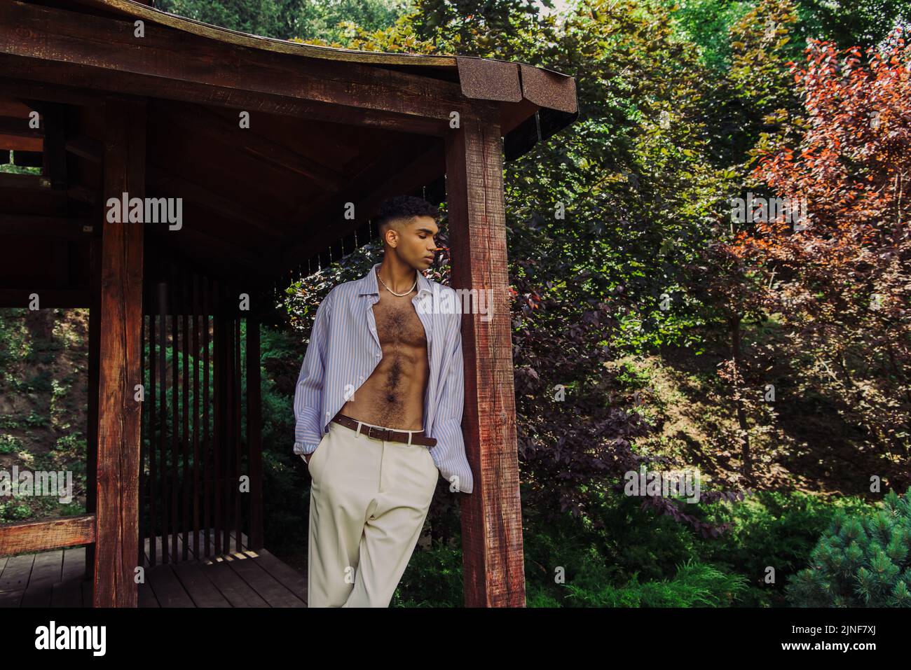 Junger afroamerikanischer Mann, der mit Händen in einer Tasche aus weißen Hosen in einem Holzpavillon im Park steht, Stockbild Stockfoto