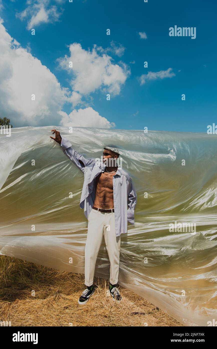 afroamerikanischer Mann in blauem Hemd und weißer Hose in der Nähe von Polyethylenfolie im Feld, Stockbild Stockfoto