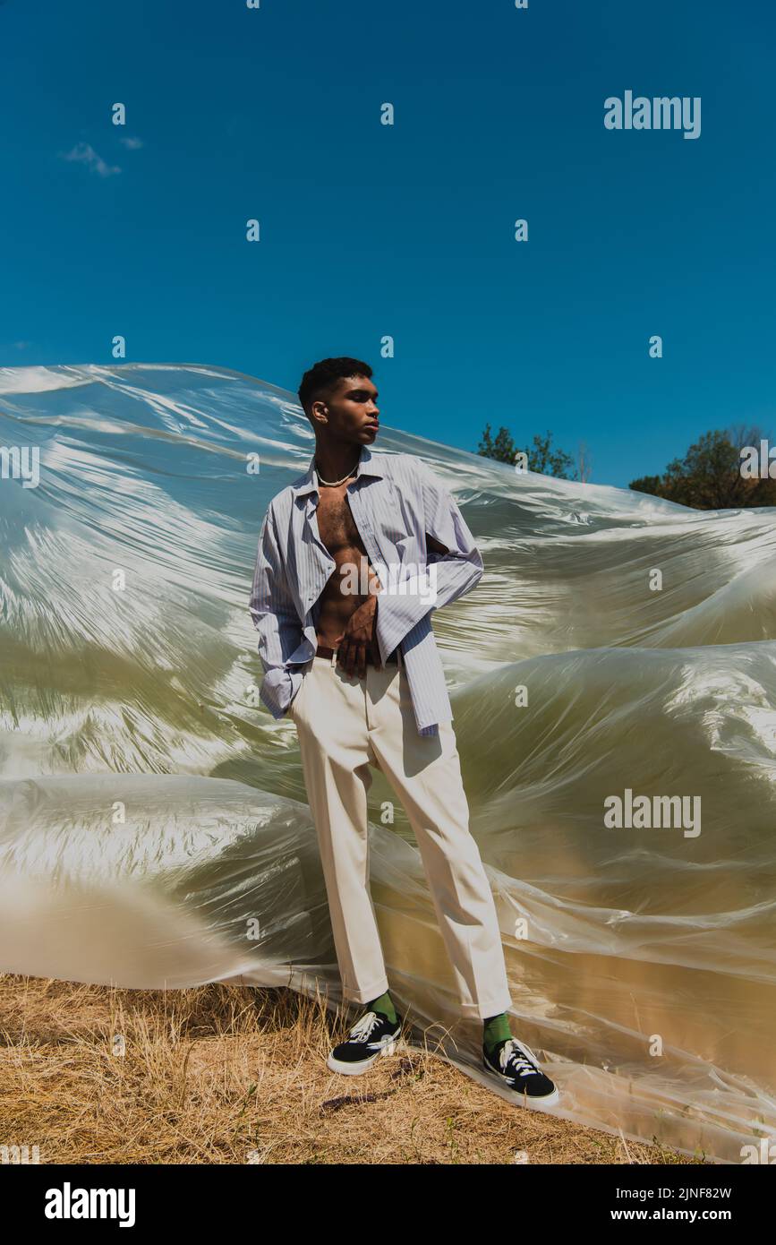 Junger afroamerikanischer Mann mit der Hand in einer Tasche aus weißen Hosen, die auf einer Wiese in der Nähe einer Polyethylenfolie stehen, Stockbild Stockfoto
