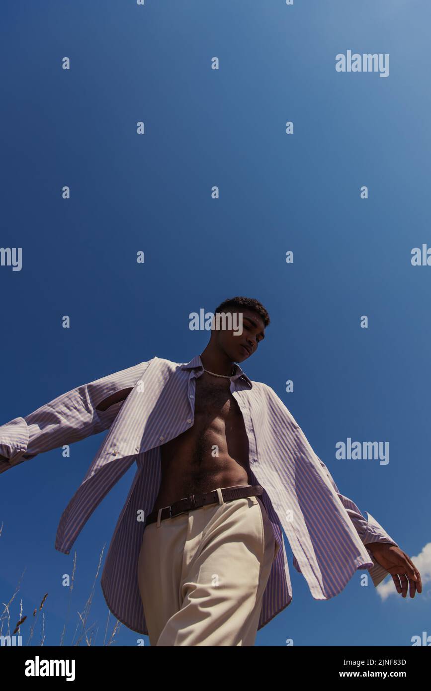 Low-Angle-Ansicht des afroamerikanischen Mannes in gestreiftem Hemd und weißer Hose gegen wolkenlosen Himmel, Stockbild Stockfoto