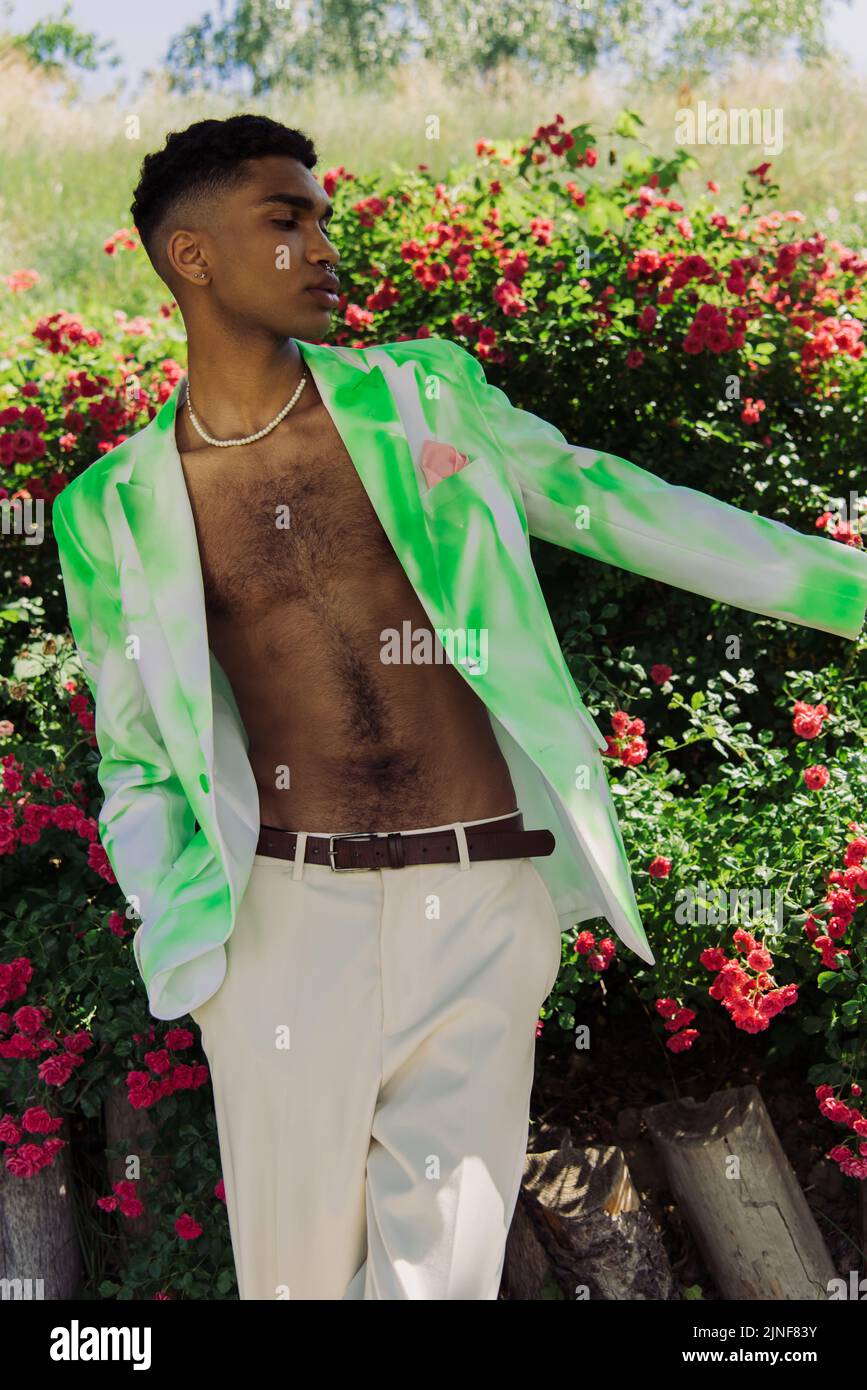 afroamerikanischer Mann in stilvollem Blazer, der mit der Hand in einer Tasche mit weißen Hosen im Park steht, Stockbild Stockfoto