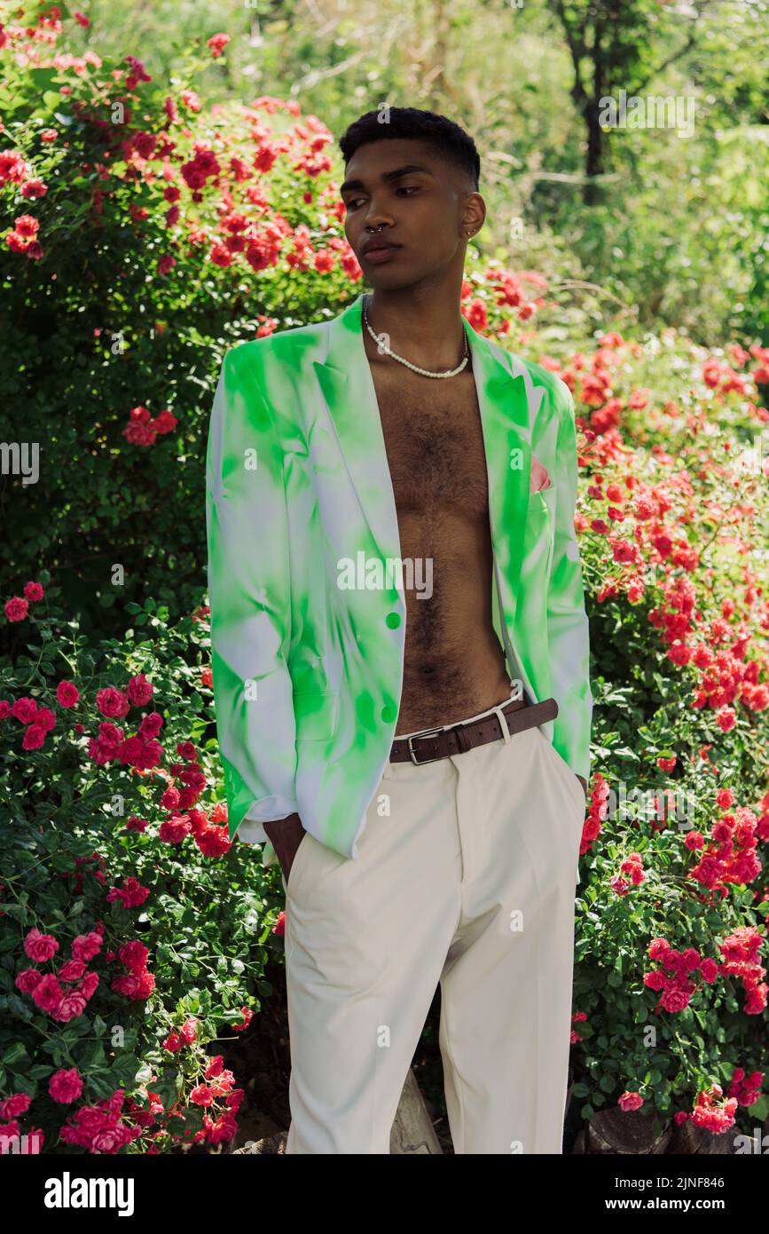 afroamerikanischer Mann in trendigem Blazer, der mit Händen in Taschen aus weißen Hosen im Park steht, Stockbild Stockfoto