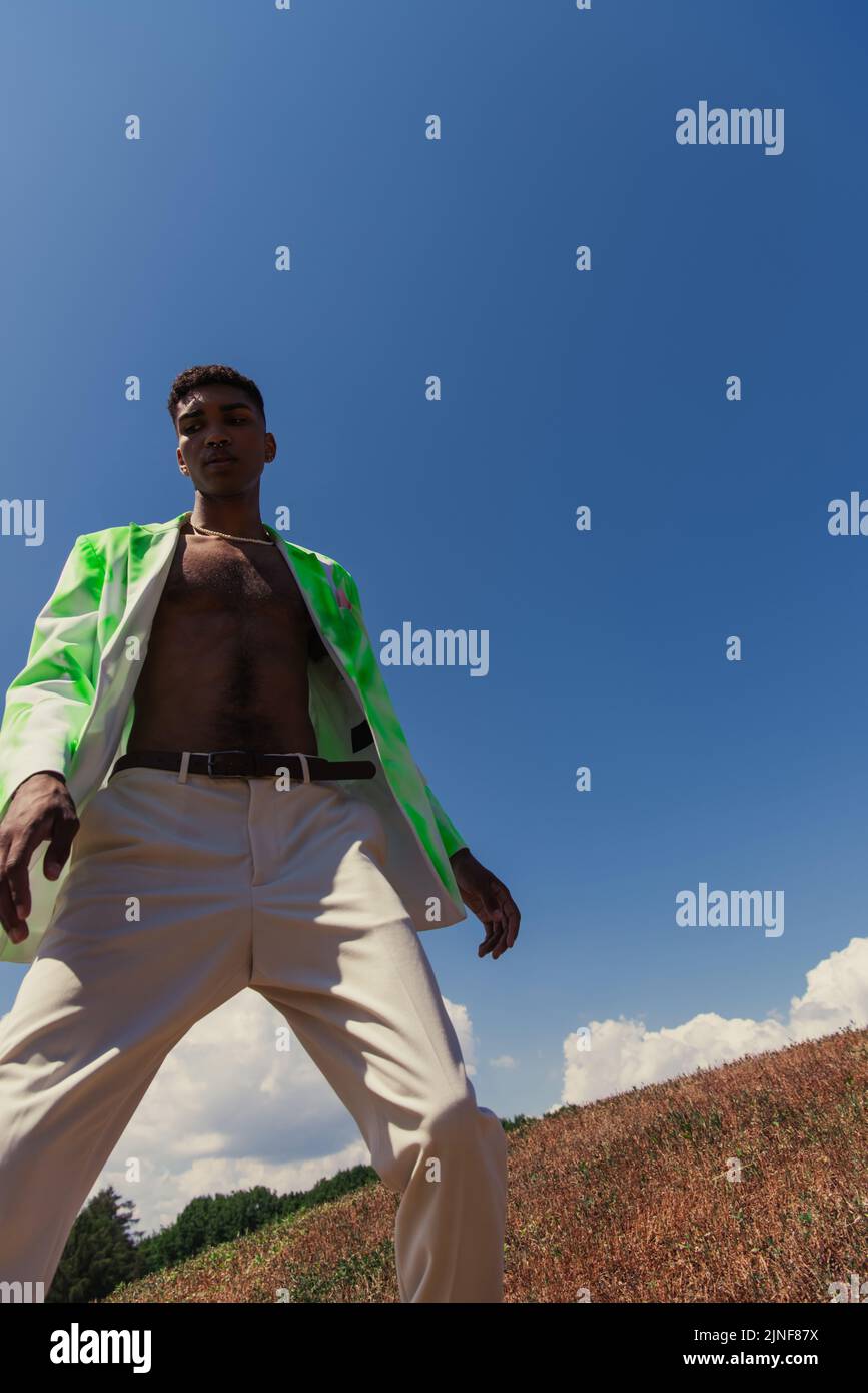 Low-Angle-Ansicht des afroamerikanischen Mannes in weißen Hosen und trendigen Blazer gegen blauen Himmel, Stockbild Stockfoto