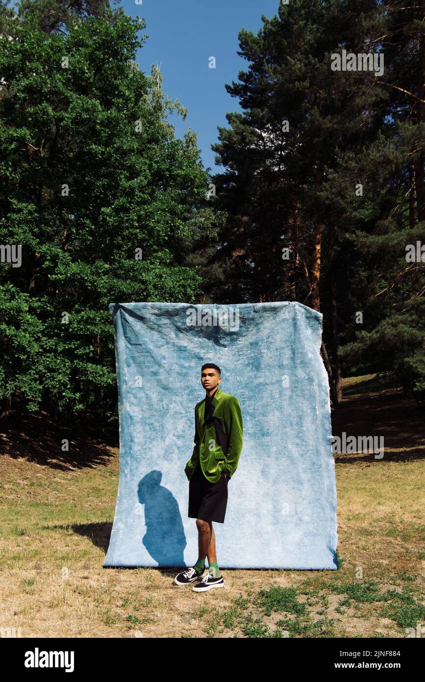 afroamerikanischer Mann in grünem Blazer posiert mit Händen in Taschen von schwarzen Shorts in der Nähe von blauem Tuch im Park, Stock Bild Stockfoto