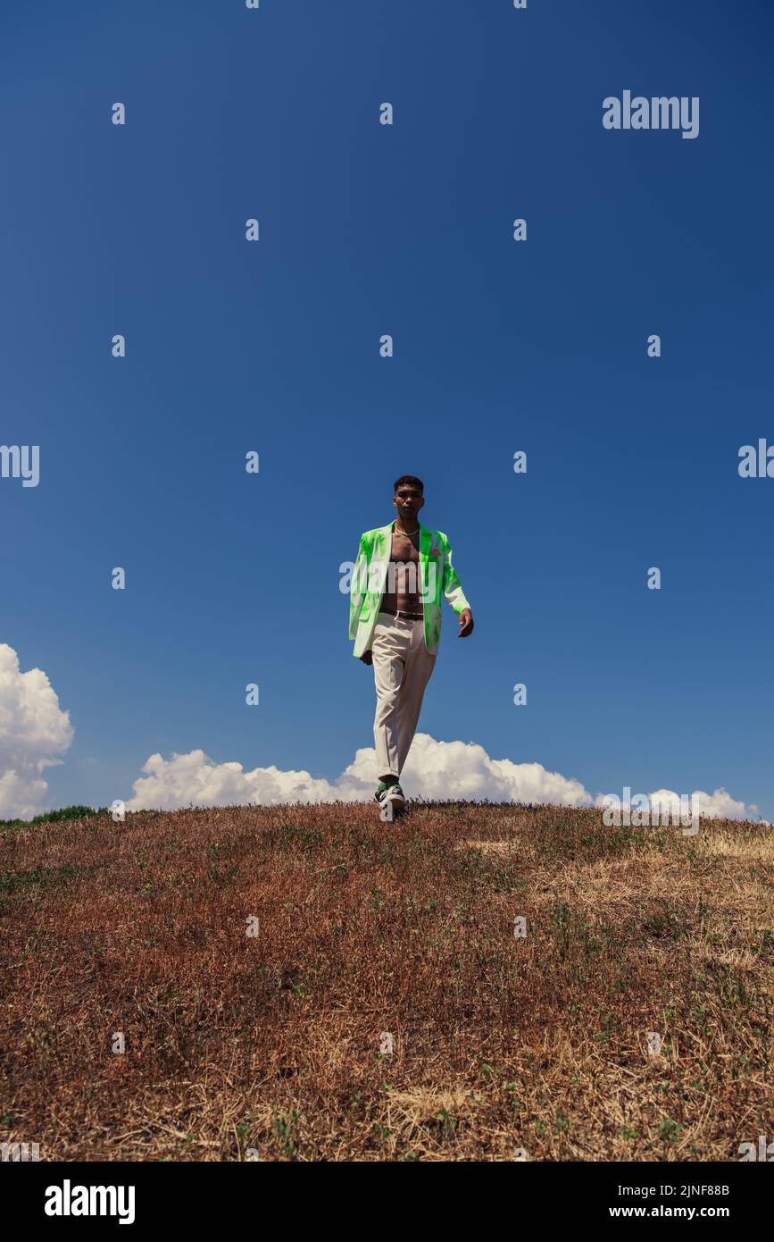 In voller Länge ein afroamerikanischer Mann in Hosen und Blazer, der unter blauem Himmel mit weißen Wolken auf dem Feld läuft, Stockbild Stockfoto