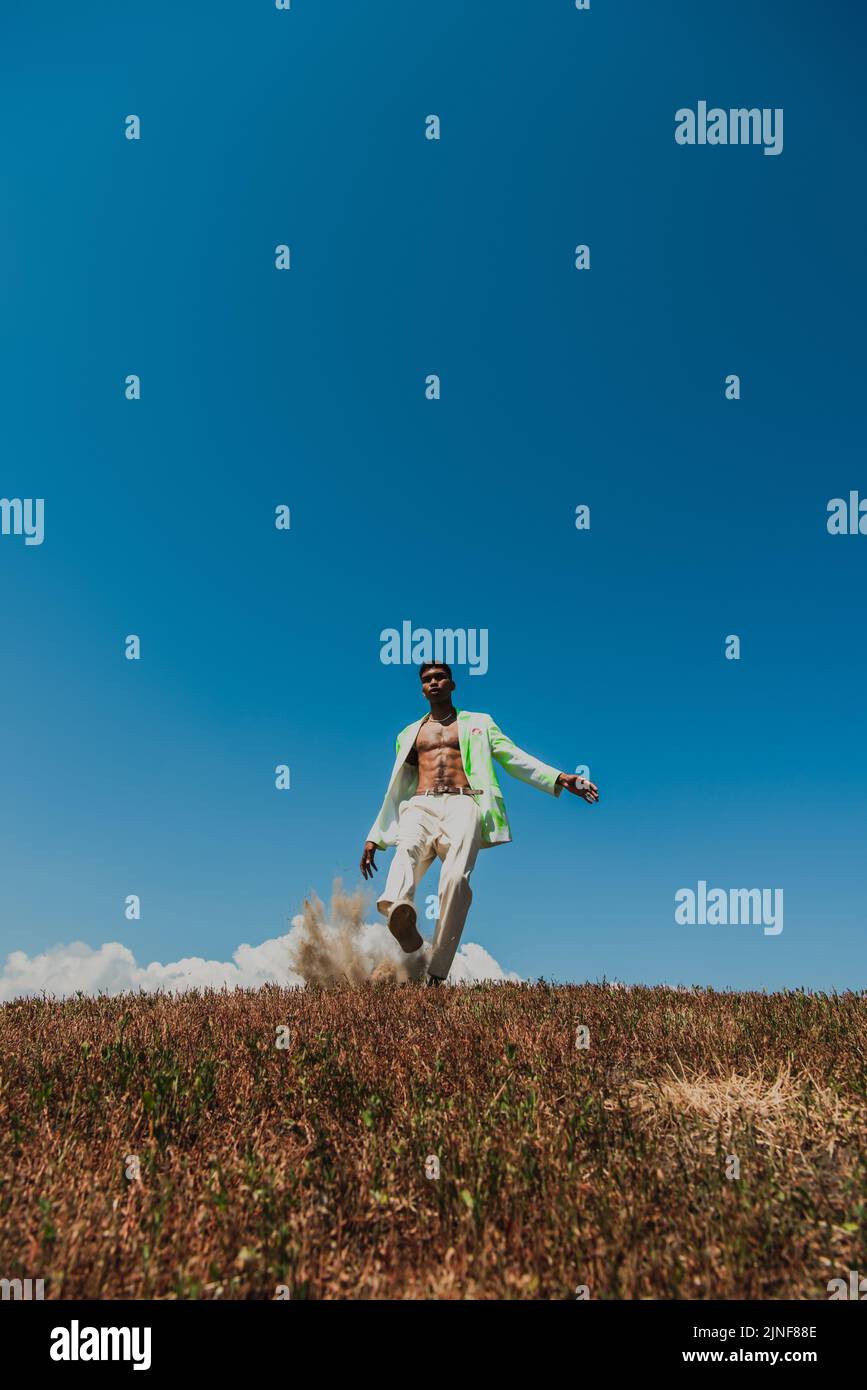 Junger afroamerikanischer Mann in weißer Hose unter blauem Himmel auf Wiese, Stockbild Stockfoto