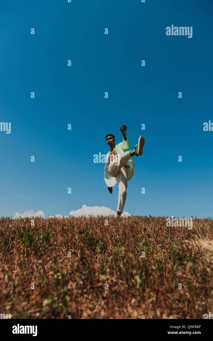 Trendiger afroamerikanischer Mann, der auf einem Bein im Grasfeld unter blauem Himmel posiert, Stockbild Stockfoto