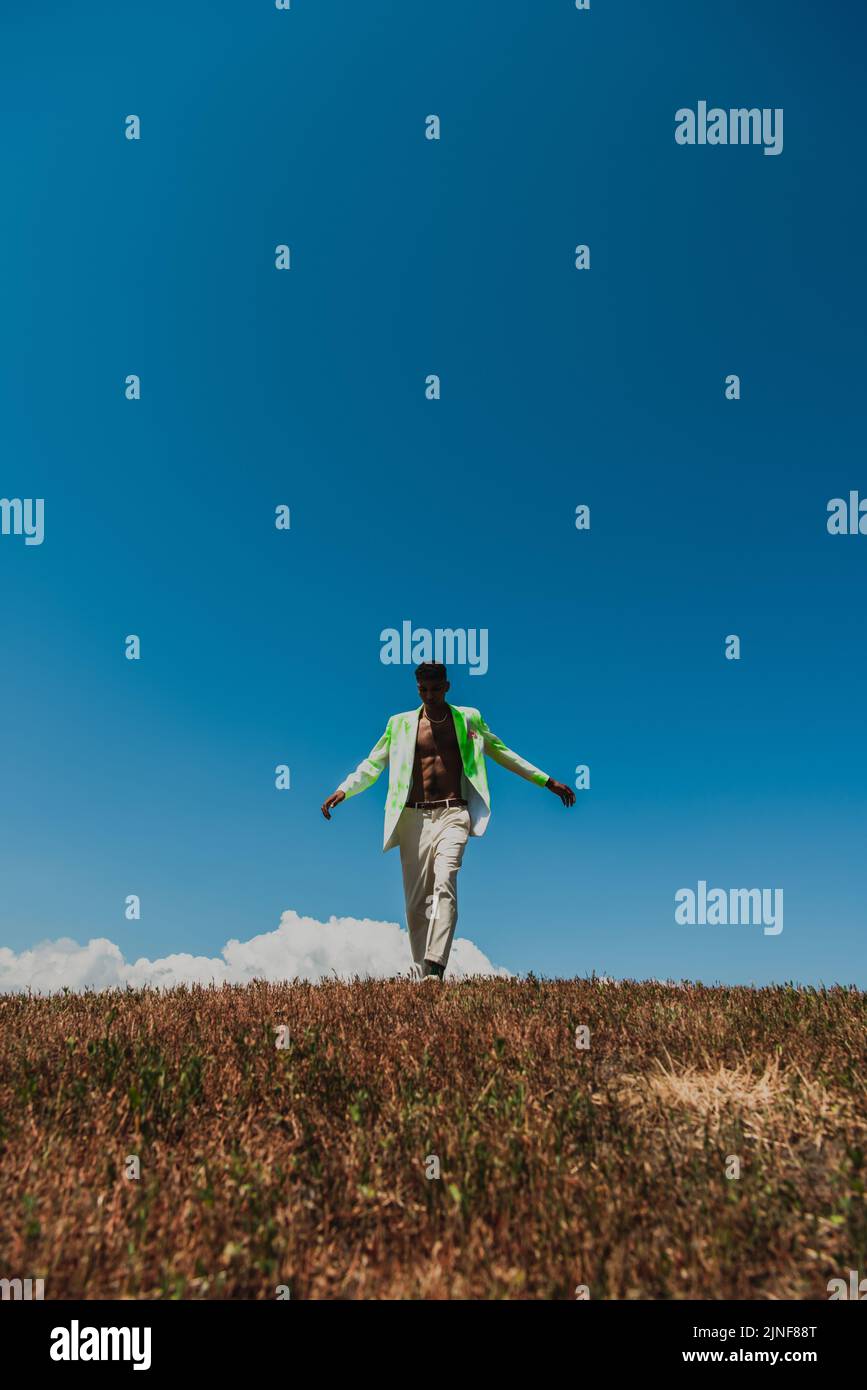 afroamerikanischer Mann in weißen Hosen und Blazer unter blauem Himmel im Feld, Stockbild Stockfoto