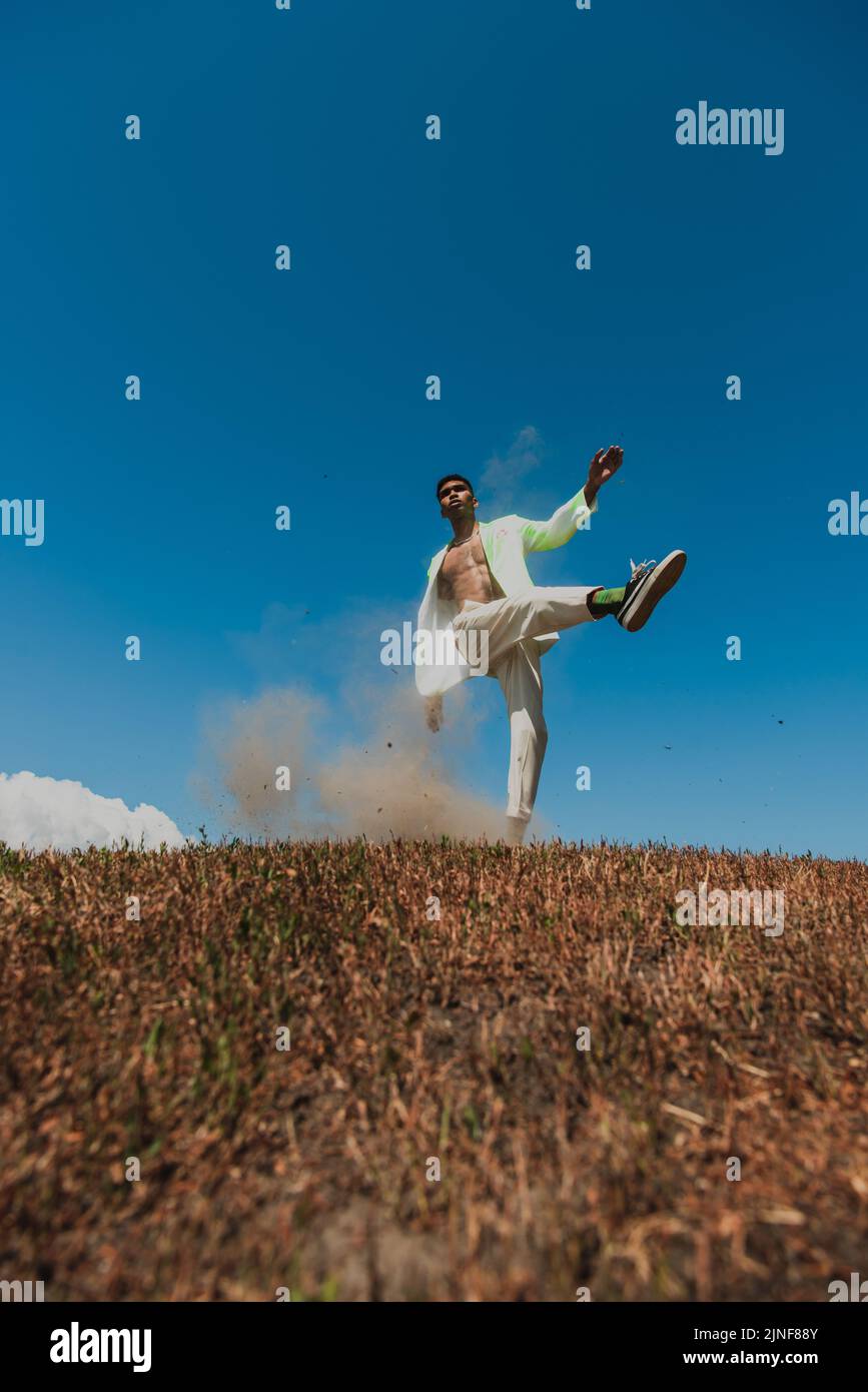 Junger und trendiger afroamerikanischer Mann, der auf einem Bein auf einem grasbewachsenen Feld unter klarem Himmel posiert, Stockbild Stockfoto