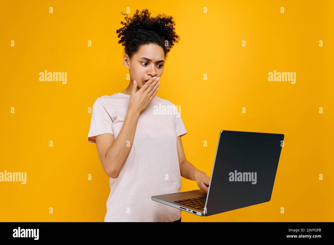 Verblüfft afroamerikanische junge Frau mit lockigen Haaren, blickt überrascht auf den Laptop-Bildschirm, sah unerwartete Nachrichten, steht über isoliertem orangefarbenem Hintergrund, emotionalem Gesichtsausdruck, der ihren Mund bedeckt Stockfoto