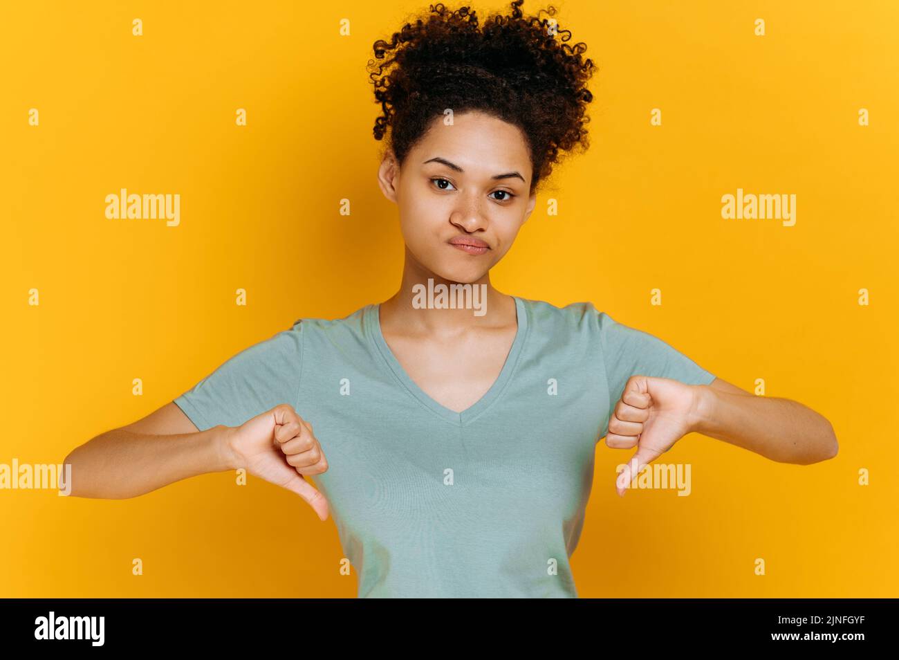 Negativ pessimistische junge afroamerikanische Frau mit lockigen Haaren, in einfachem T-Shirt gekleidet, zeigt Daumen nach unten Geste, blickt auf die Kamera, stehend auf isoliertem orangefarbenem Hintergrund, trauriger Gesichtsausdruck Stockfoto