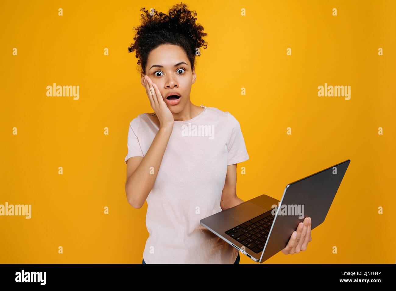 Schockierte afroamerikanische junge Frau mit lockigen Haaren, hält einen offenen Laptop in der Hand, schaut in atemberaubender Form auf die Kamera, erhält unerwartete Informationen, steht über isoliertem orangefarbenem Hintergrund, emotionaler Ausdruck Stockfoto