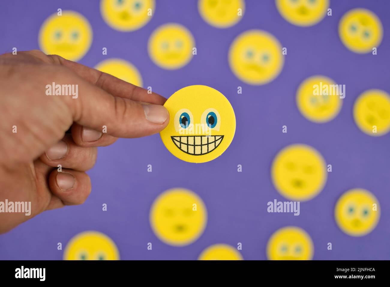Nahaufnahme einer Hand, die ein gelbes Smiley-Gesicht Emoji zwischen traurigen Emojis auf lila Hintergrund hält Stockfoto