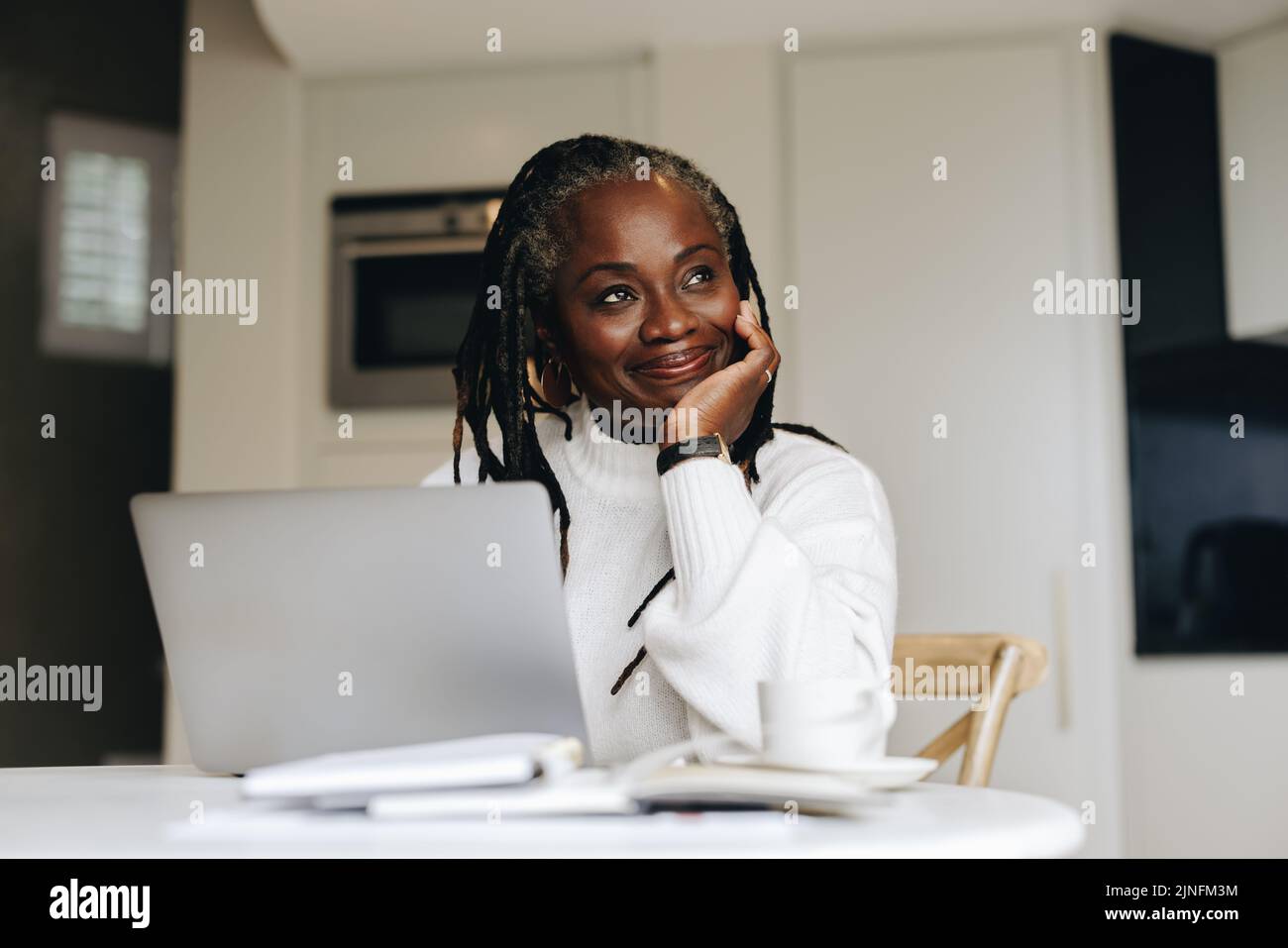 Eine fröhliche Geschäftsfrau, die zu Hause am Laptop arbeitet, schaut nachdenklich weg. Reife Geschäftsfrau macht online freiberufliche Arbeit in ihrem hom Stockfoto