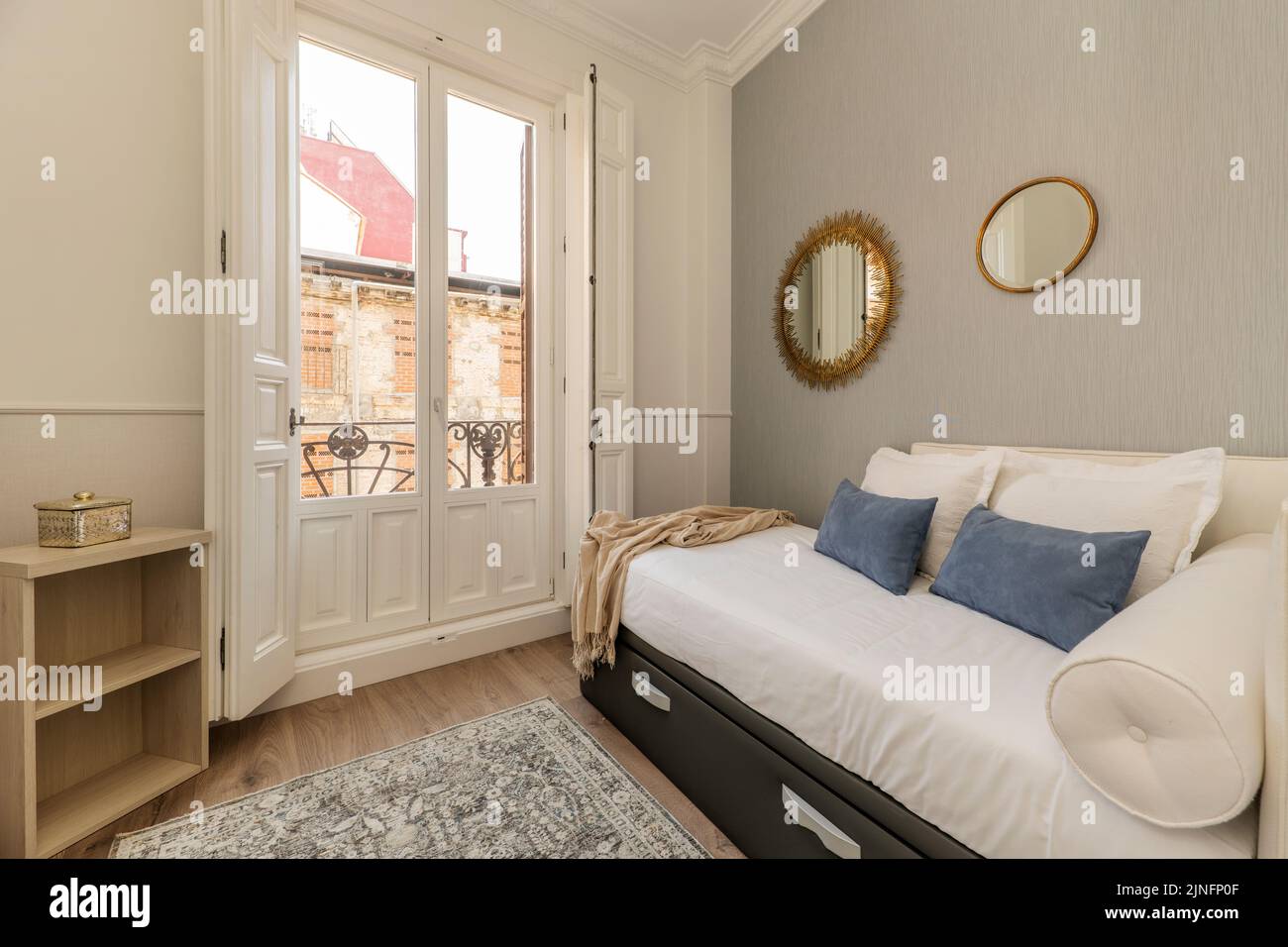 Schlafzimmer mit Einzelbett mit gepolsterter ausziehbarer Kommode, weißen und blauen Kissen und dekorativen Spiegeln neben weißem Balkon mit Fenstern Stockfoto