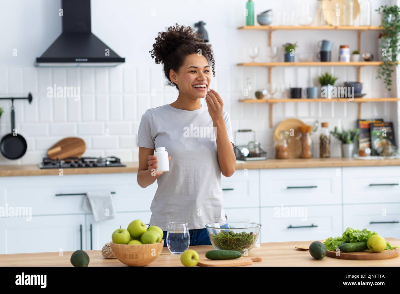 Glückliche afroamerikanische Frau, die am Küchentisch in der heimischen Küche steht und Nahrungsergänzungsmittel trinkt Stockfoto