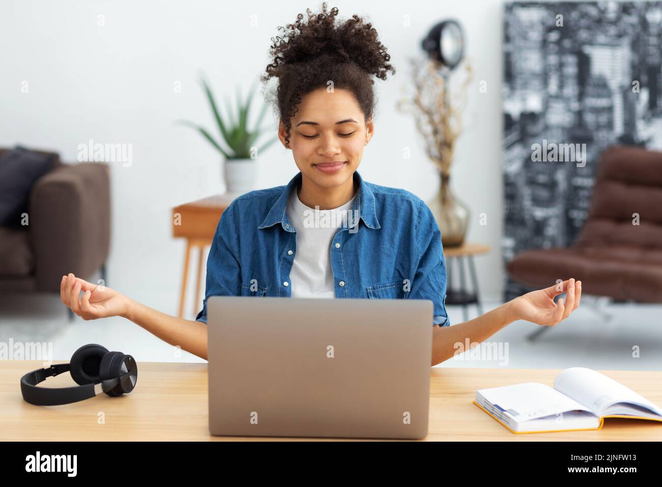 Junge afroamerikanische Frau, die mit einem Laptop am Schreibtisch sitzt und die Augen geschlossen meditiert und an gute Dinge denkt Stockfoto