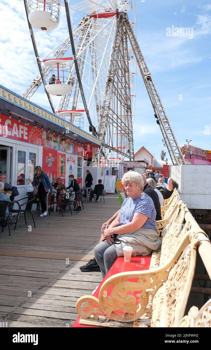 Blackpool, Lancashire, Großbritannien August 6 2022 eine ältere Dame ruht auf einer Pier-Bank mit einem großen Festplatz-Rad im Hintergrund Stockfoto Blackpool, Lancashire, Großbritannien August 6 2022 eine ältere Dame ruht auf einer Pier-Bank mit einem großen Festplatz-Rad im Hintergrund Stockfoto