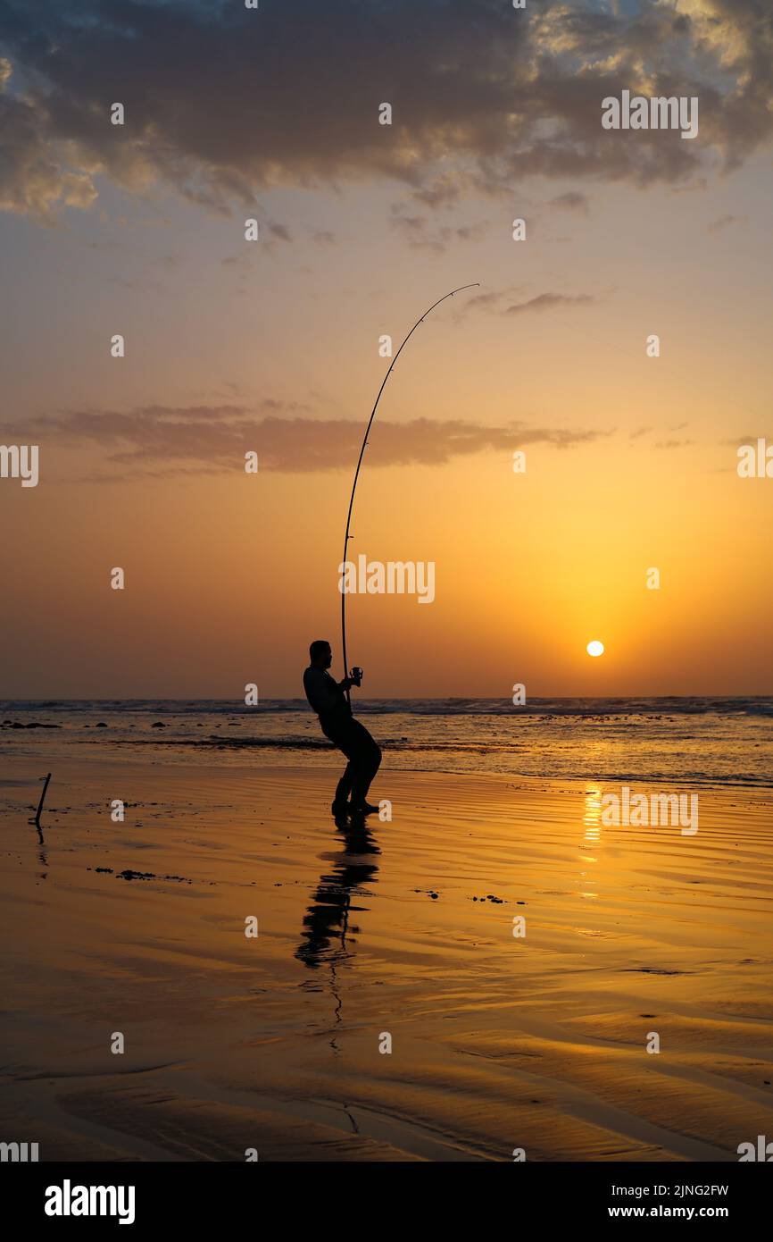 Ein Fischer, der seinen Haken mit seiner Angelrute hält, bei einem wunderschönen Sonnenuntergang in der Stadt Laayoune Stockfoto