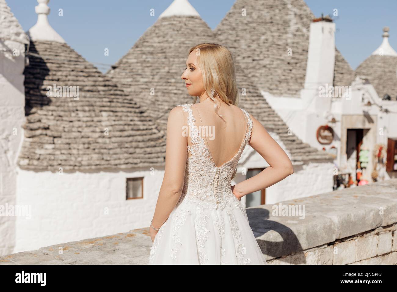 Rückansicht der jungen glücklichen Frau Braut, die in der Nähe von konischen Dächern von Trullo im sonnigen Italien steht und die Hand auf die Taille posiert. Stockfoto