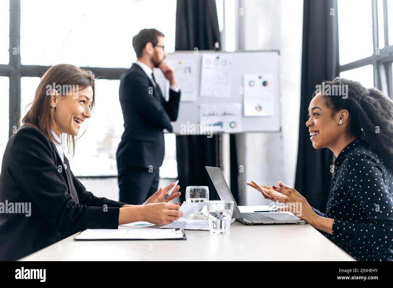 Zwei erfolgreiche, selbstbewusste Frauen verhandeln, diskutieren Geschäftsprojekt, planen Finanzstrategie, lächeln. Eleganter Mann steht auf dem Hintergrund in der Nähe von Whiteboard und denkt Businessplan Stockfoto