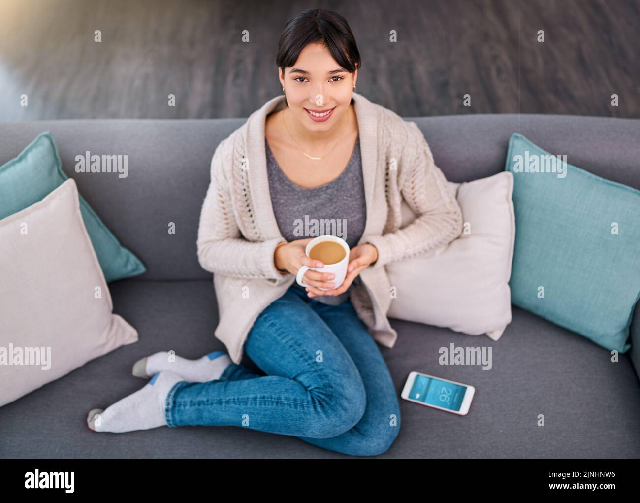 Machte eine frische Kanne Tee. Porträt einer fröhlichen jungen Frau, die tagsüber auf einem Sofa sitzt, während sie zu Hause Kaffee trinkt. Stockfoto