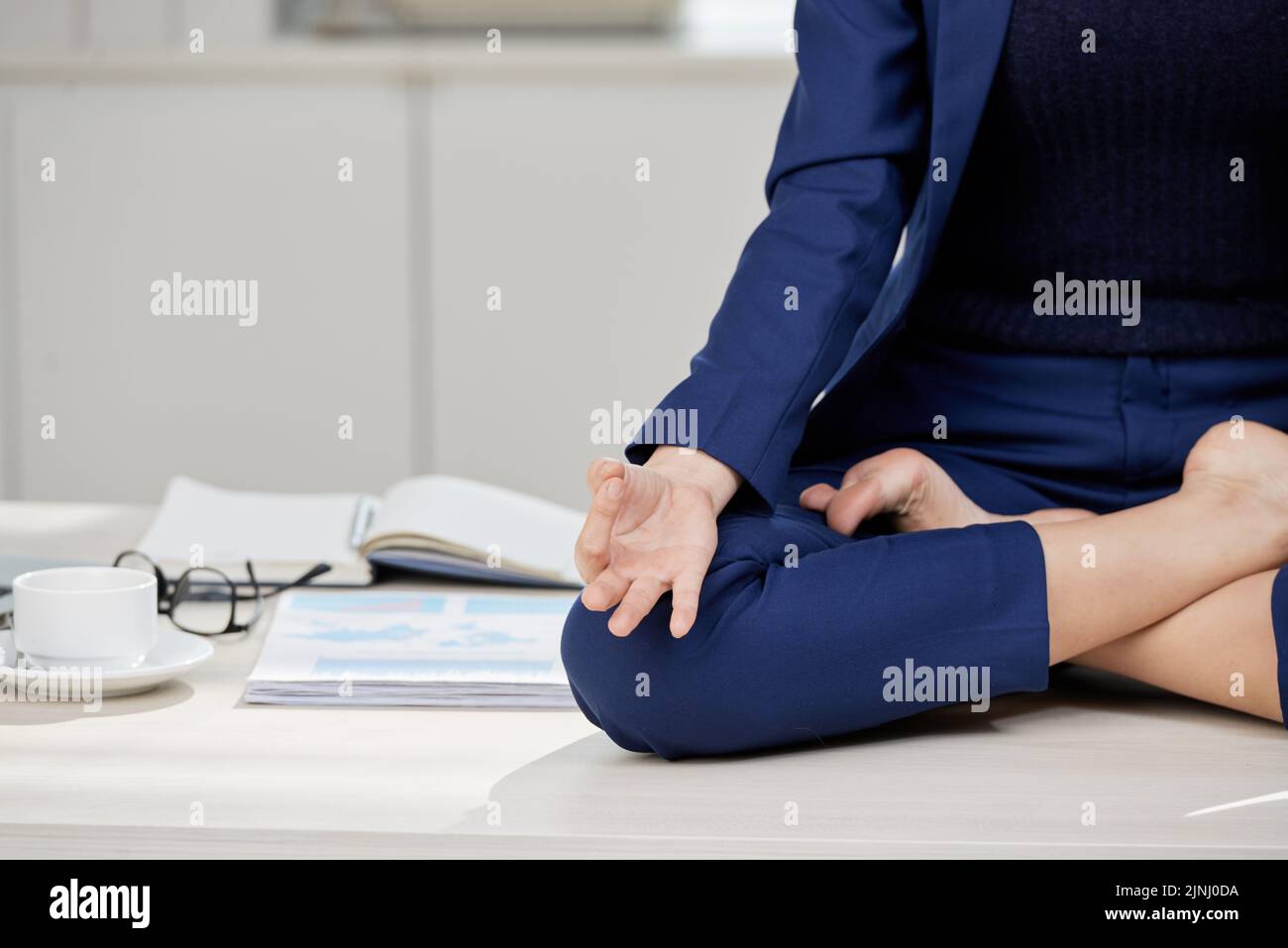 Nahaufnahme eines nicht erkennbaren jungen Managers in klassischem Anzug, der in Lotusposition auf dem Schreibtisch sitzt und Meditation praktiziert Stockfoto