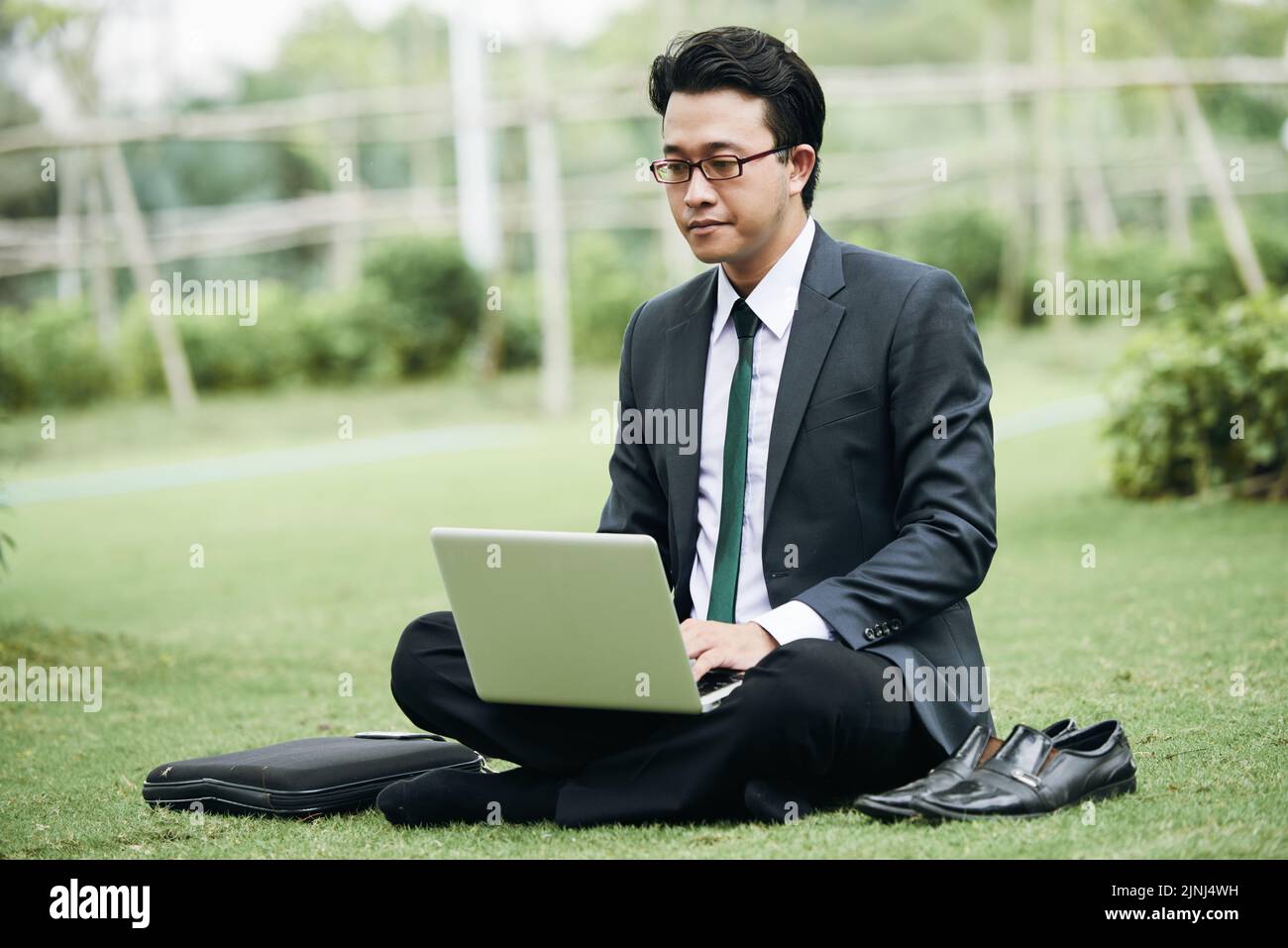 Asiatischer Geschäftsmann sitzt mit gekreuzten Beinen auf grünem Rasen und arbeitet auf einem Laptop Stockfoto