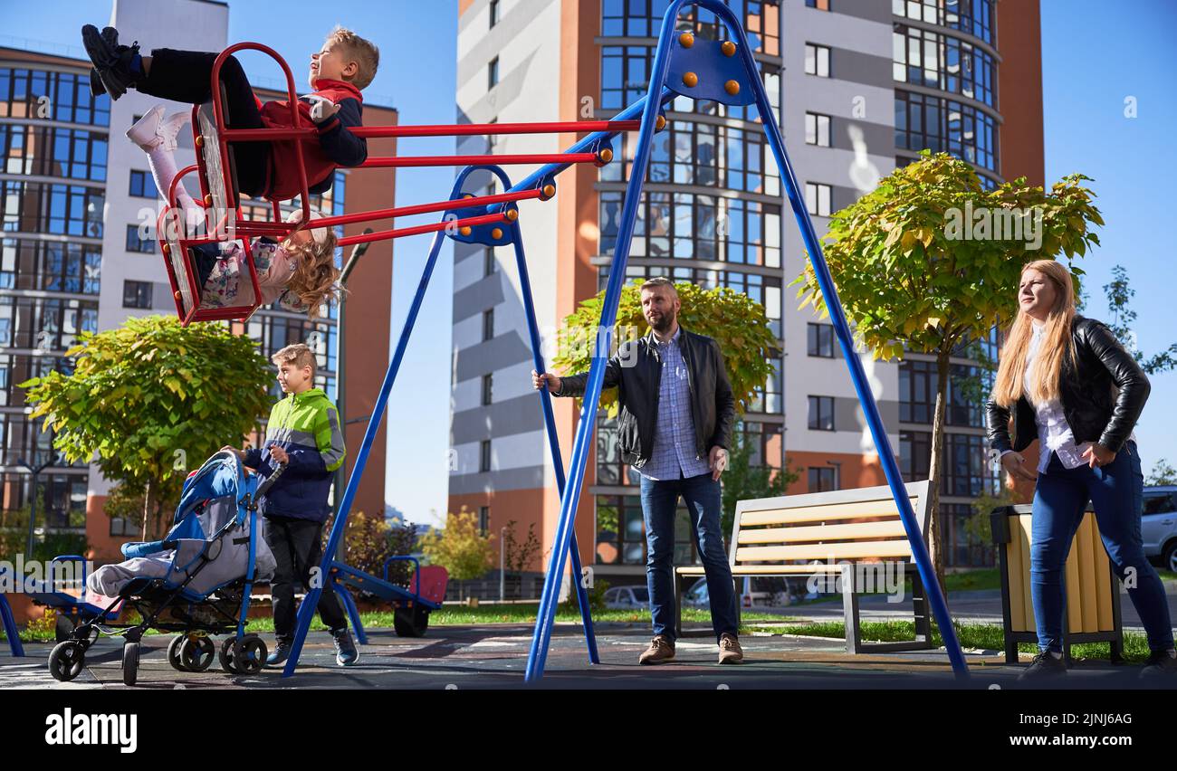 Glückliche Familie - Vater, Mutter und Kinder, die gemeinsam auf dem Spielplatz Spaß haben. Eltern schwingen Tochter und Sohn, während ein anderes Kind mit Kinderwagen steht. Moderne Wohngebäude im Hintergrund. Stockfoto