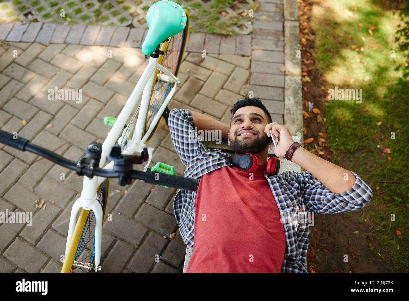 Aus dem Hochwinkel betrachtet, wie ein fröhlicher Inder auf dem Smartphone mit einem Freund spricht, während er sich vom Radfahren ausruhte, er auf einer Parkbank lag, während das Fahrrad neben ihm stand Stockfoto