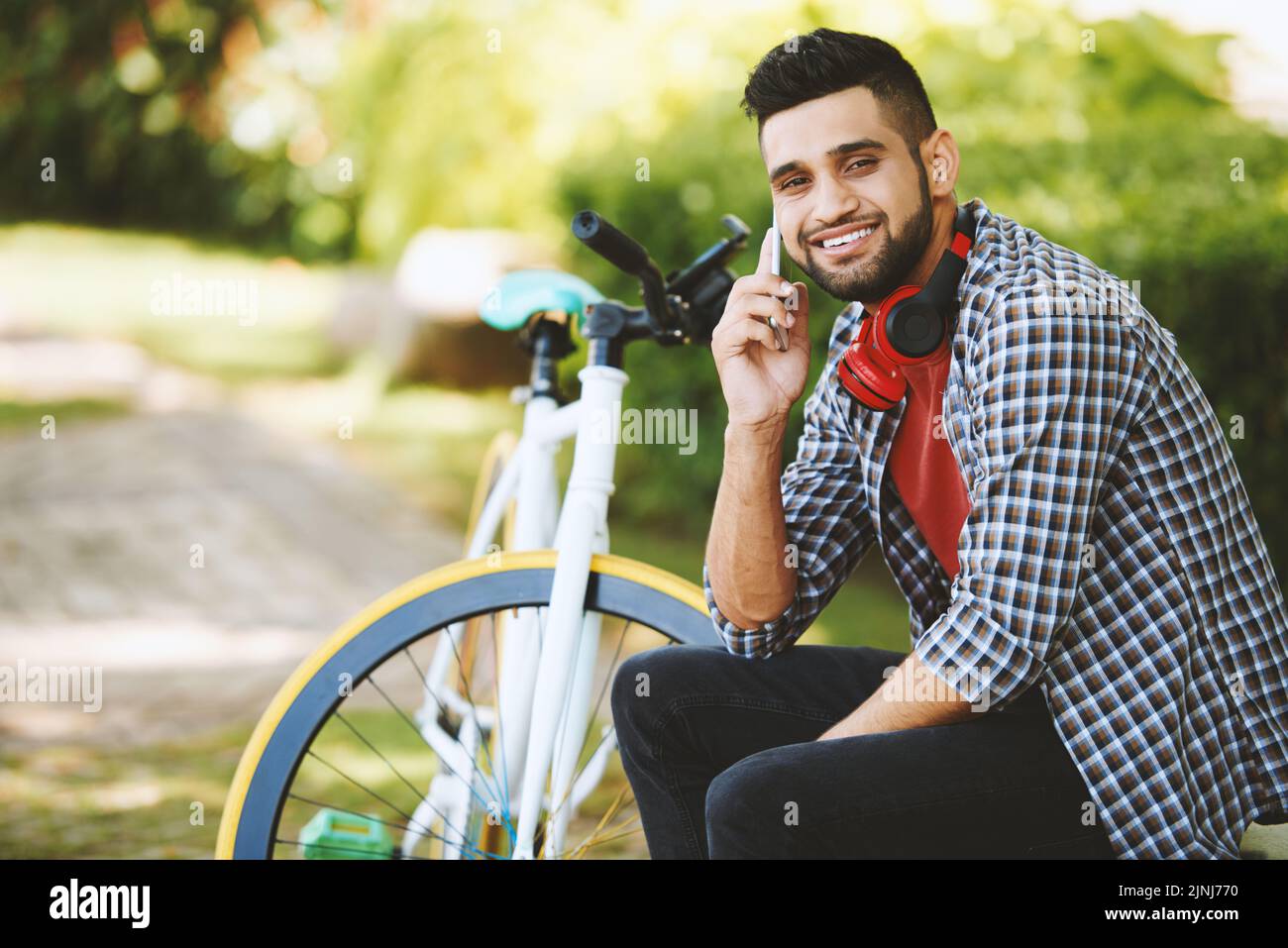 Fröhlicher indischer Mann, der mit toothy Lächeln auf die Kamera schaut, während er mit einem Freund auf dem Smartphone spricht und auf einer Parkbank sitzt, modernes Fahrrad im Hintergrund Stockfoto