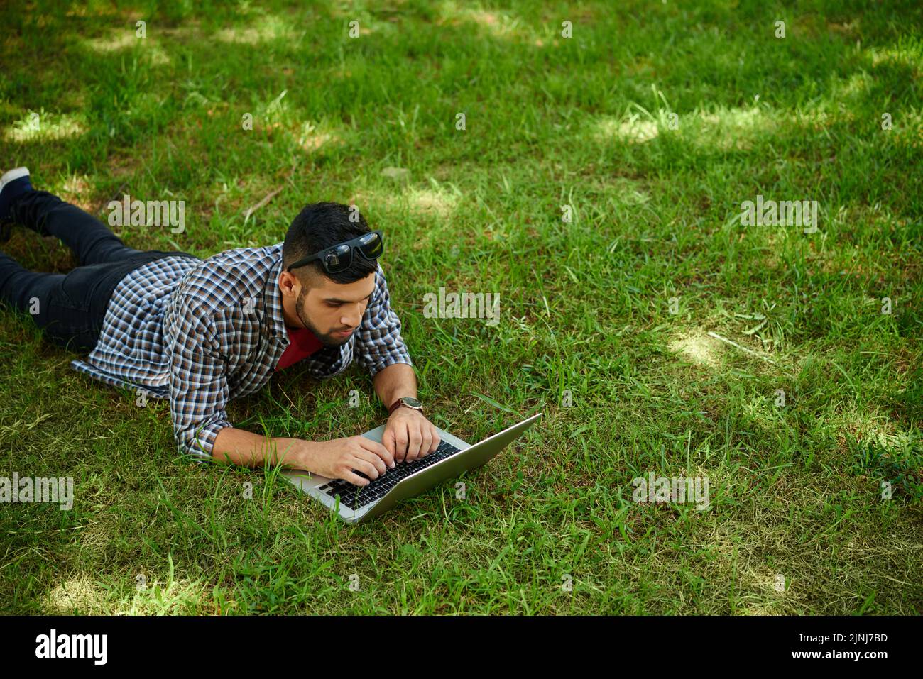 Gutaussehender bärtiger Designer in Casualwear, der auf grünem Rasen liegt, während er mit Hilfe eines Laptops an einem Start-up-Projekt arbeitete, Portraitaufnahme Stockfoto