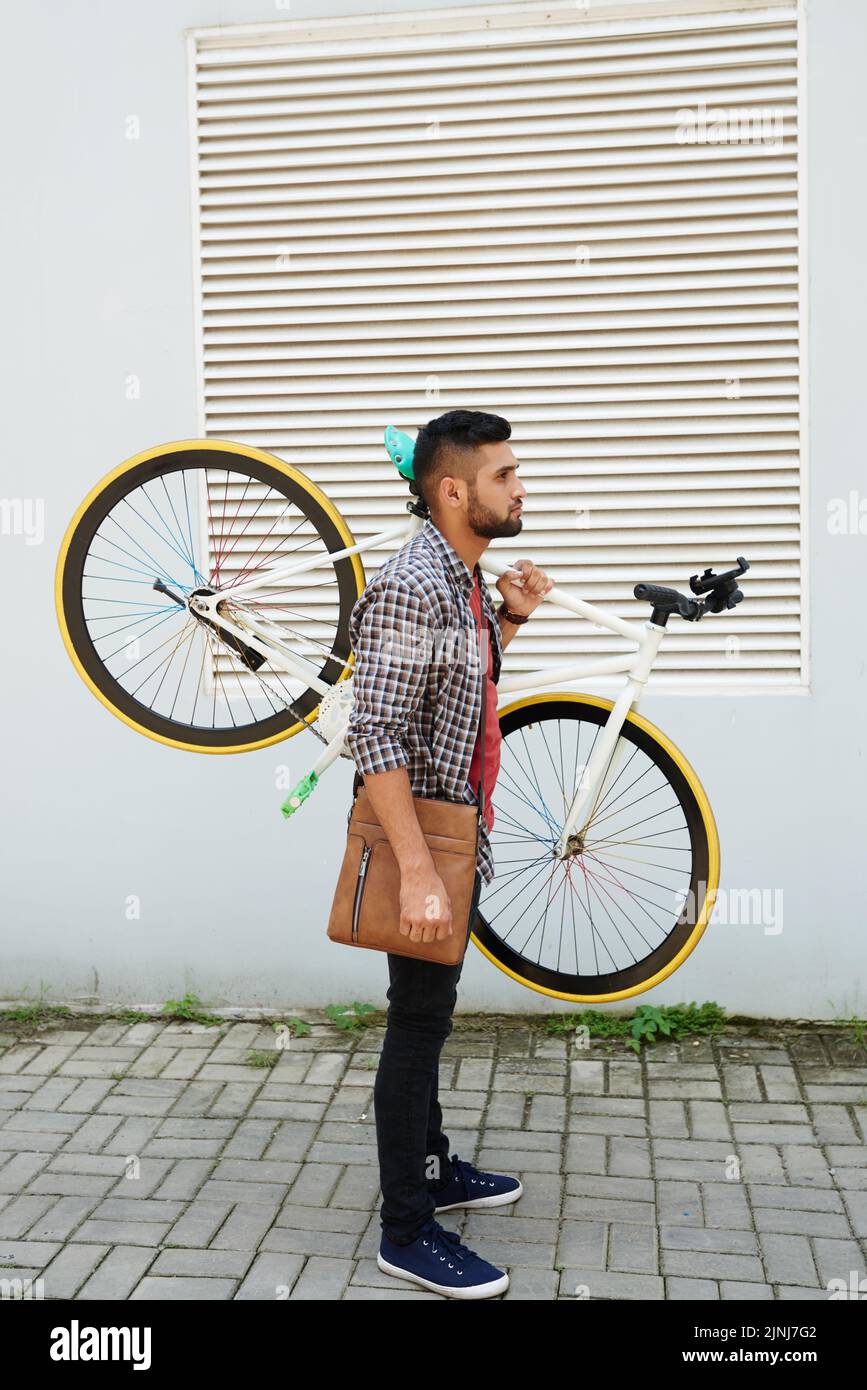 Profilansicht eines bärtigen jungen Mannes in Casualwear, der ein modernes Fahrrad auf der Schulter hält, während er an der Gebäudefassade steht, Ganzkörperportrait Stockfoto