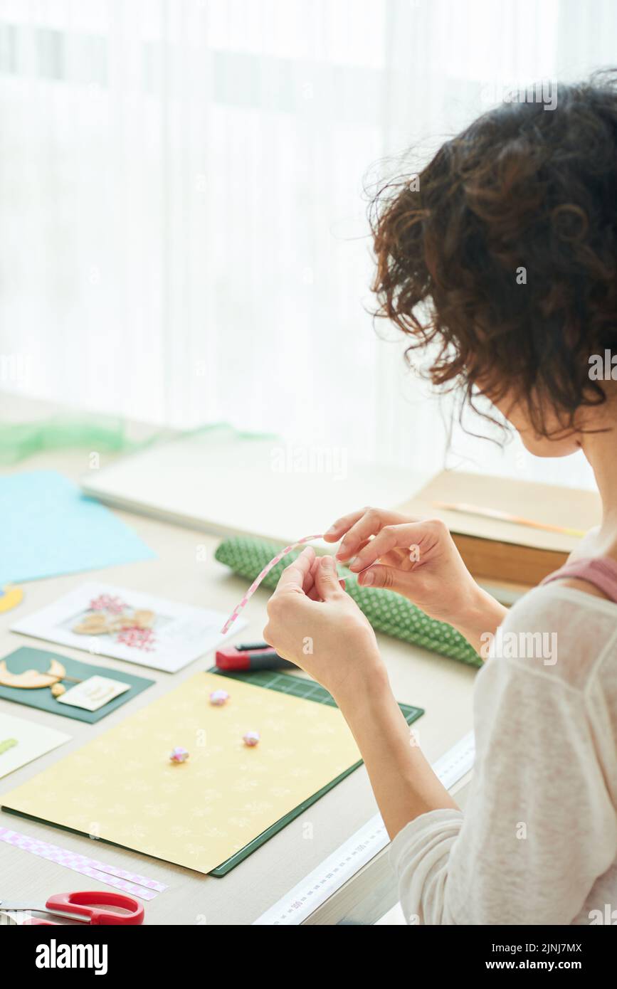 Profilansicht einer lockigen Frau in Casualwear, die am Holztisch sitzt und dabei den Prozess der handgefertigten Erstellung von Grußkarten genießt Stockfoto