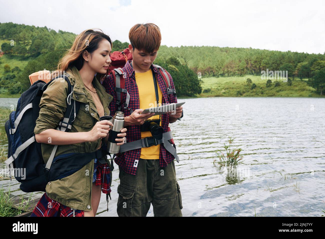 Liebevolles asiatisches Paar studiert Karte auf modernem digitalem Tablet, während Pause vom Wandern, hübsches junges Mädchen hält Vakuumflasche in den Händen, malerischer Blick auf Hintergrund Stockfoto
