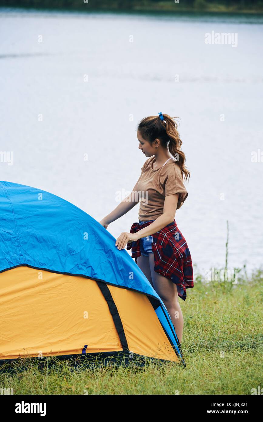 Selbstbewusste junge Frau, die T-Shirt und Shorts trägt und Zelt aufstellt, während sie beim Wandern Urlaub macht, Wasseroberfläche auf dem Hintergrund Stockfoto