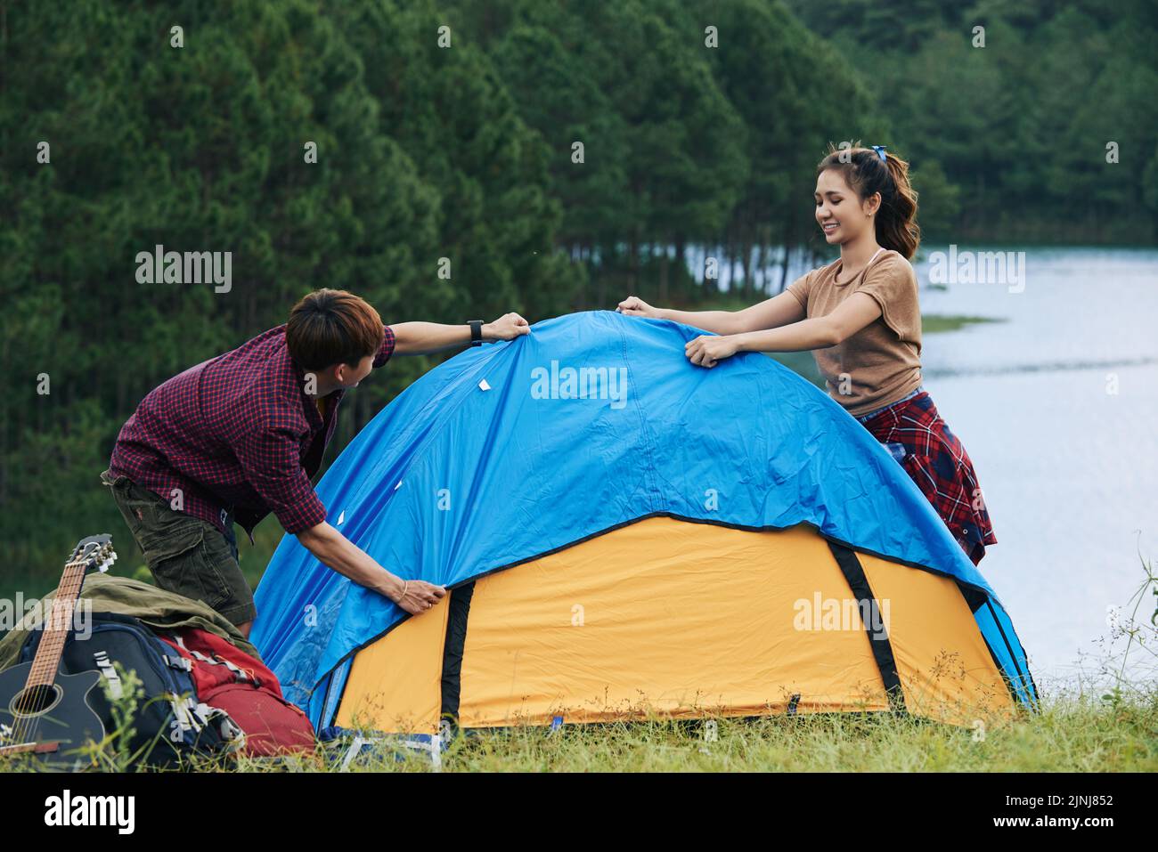 Fröhliche asiatische Touristen haben Spaß beim Aufstellen Zelt in malerischen Ort, grünen Wald und tiefen See im Hintergrund Stockfoto