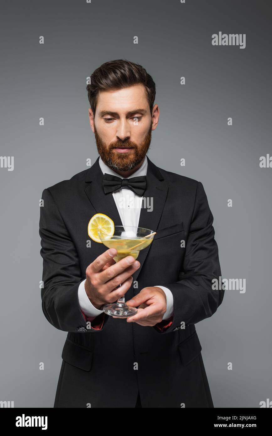 Bärtiger Mann in elegantem Anzug mit Fliege hält Glas mit Cocktail isoliert auf grau, Stock Bild Stockfoto