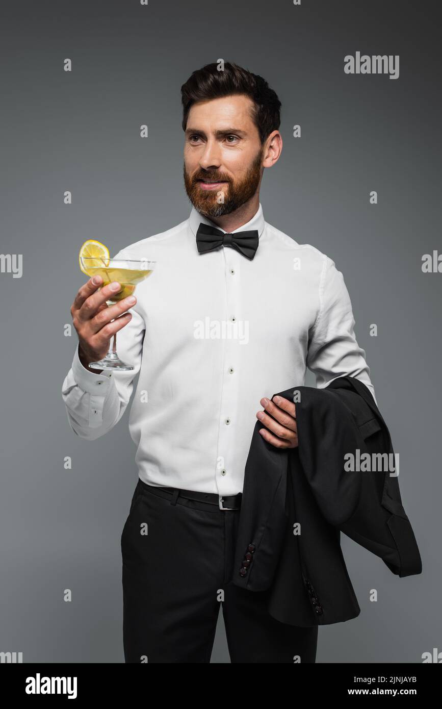 Bärtiger Mann im Smoking mit Fliege, der Glas mit Cocktail und Blazer auf grauem, Stockbild hält Stockfoto