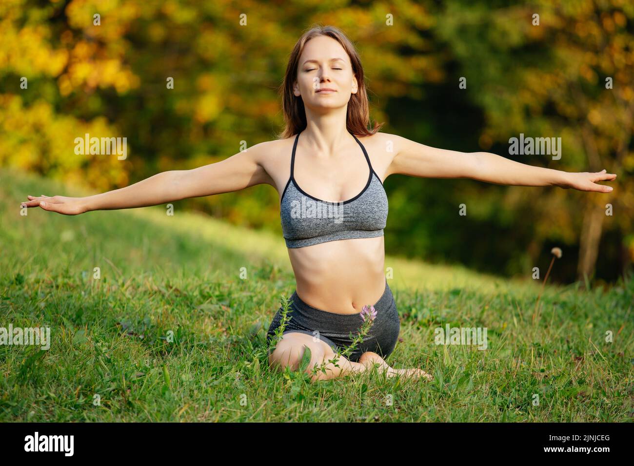 Junge Frau in Sportkleidung, die Yoga und Meditation praktiziert und draußen vor der Stadt Atemübungen macht. Frau trainiert morgens auf dem Gras Stockfoto